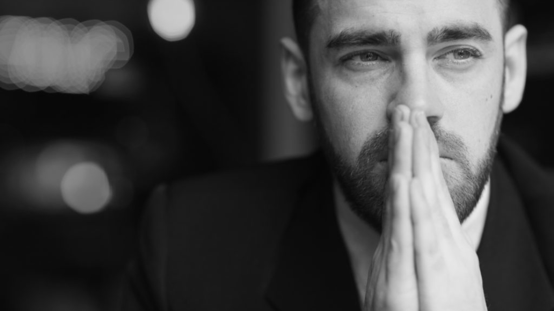 Portrait of modern bearded businessman looking away with red teary eyes, stressed and tired against black background, covering his mouth with hands